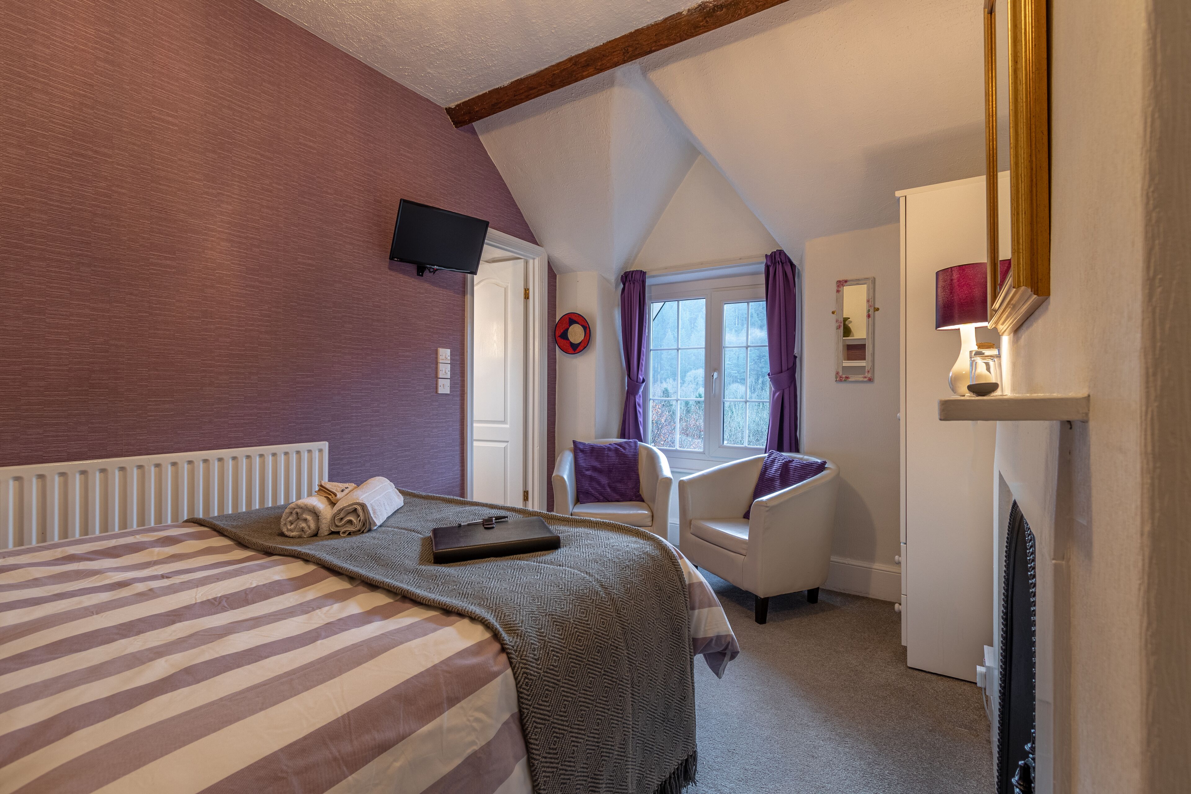 A hotel room with a bed, a chair, a TV, a window with curtains, and a fireplace.