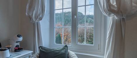 A room with a window, a bed, a bedside table with a lamp, and a view of trees outside.