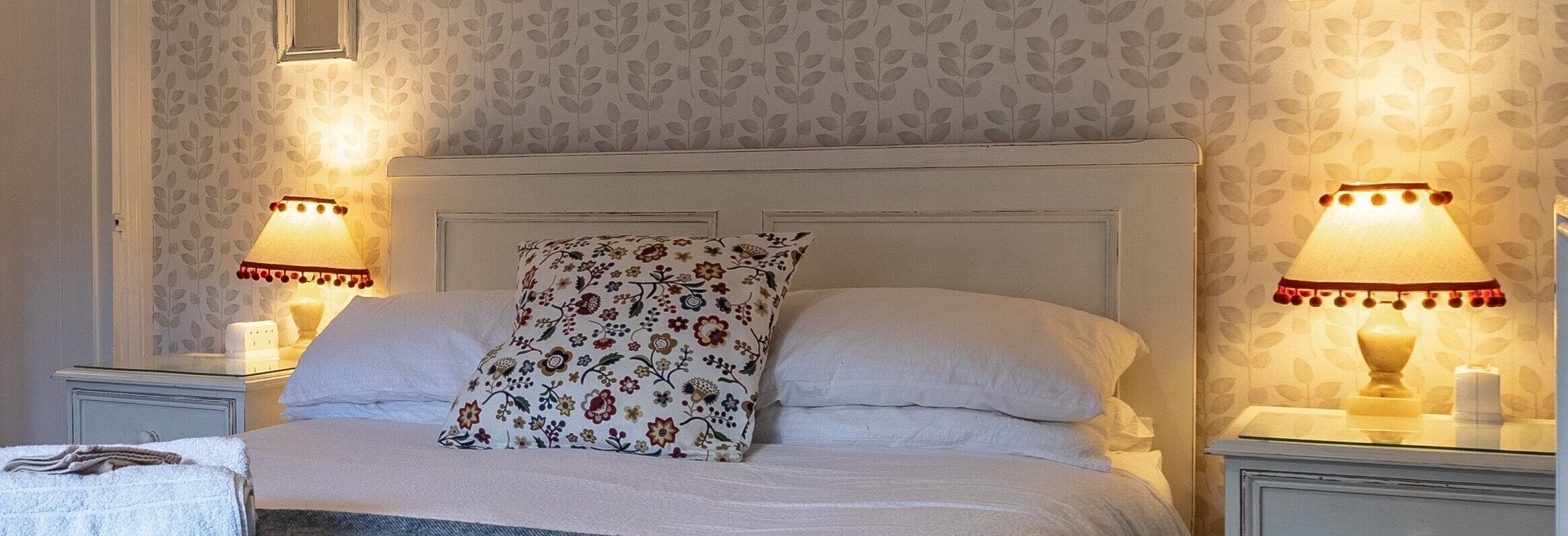 Un lit bien fait, agrémenté de coussins à motifs floraux, de deux lampes de chevet et d'un papier peint à motifs.