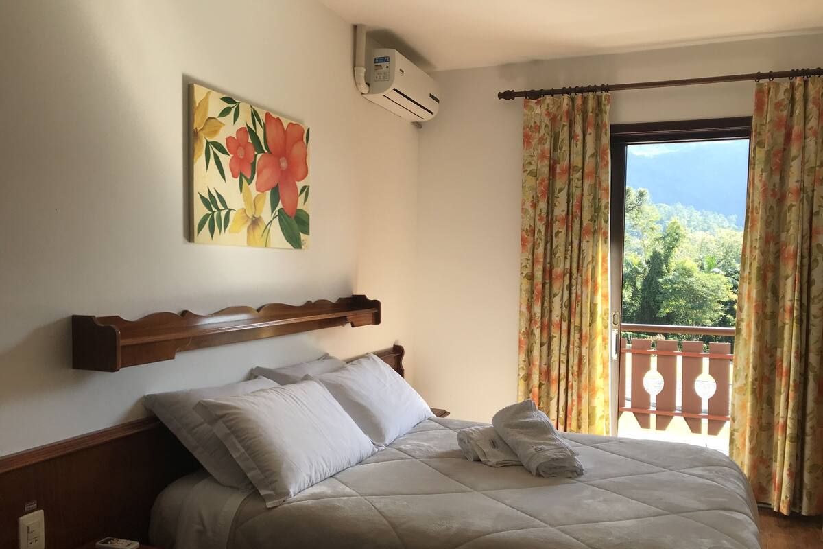 A bedroom with a bed, a painting, an air conditioner, and a view of greenery outside.