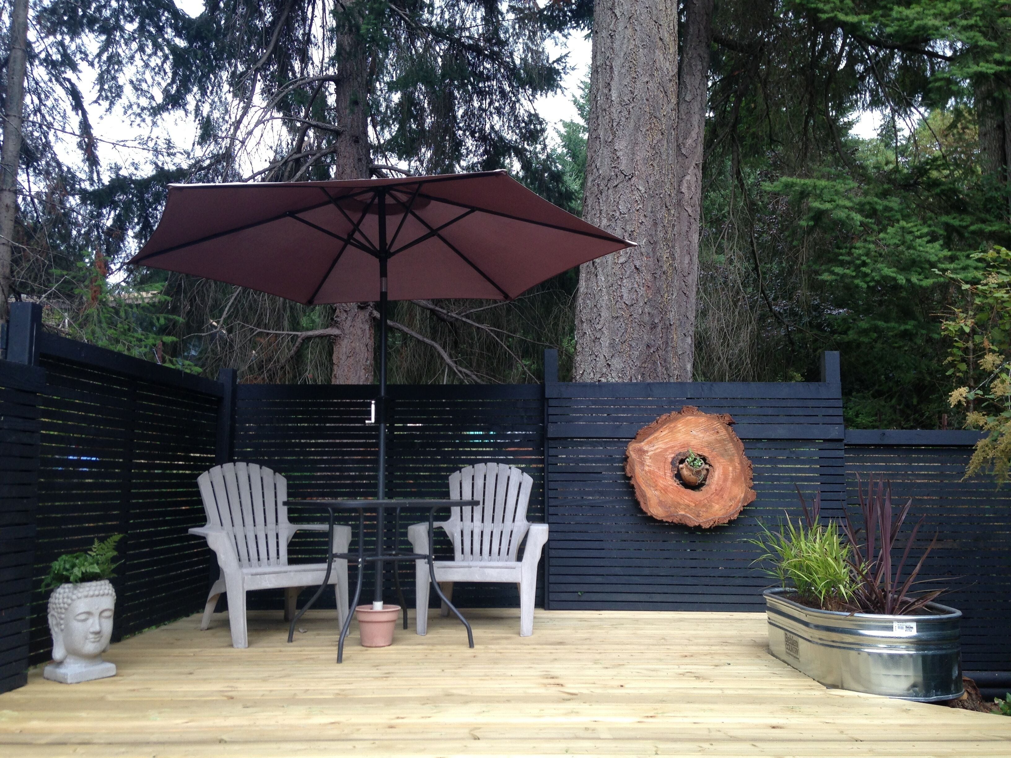 An outdoor patio with a table and two chairs, a large umbrella, and a decorative tree ring wall hanging.