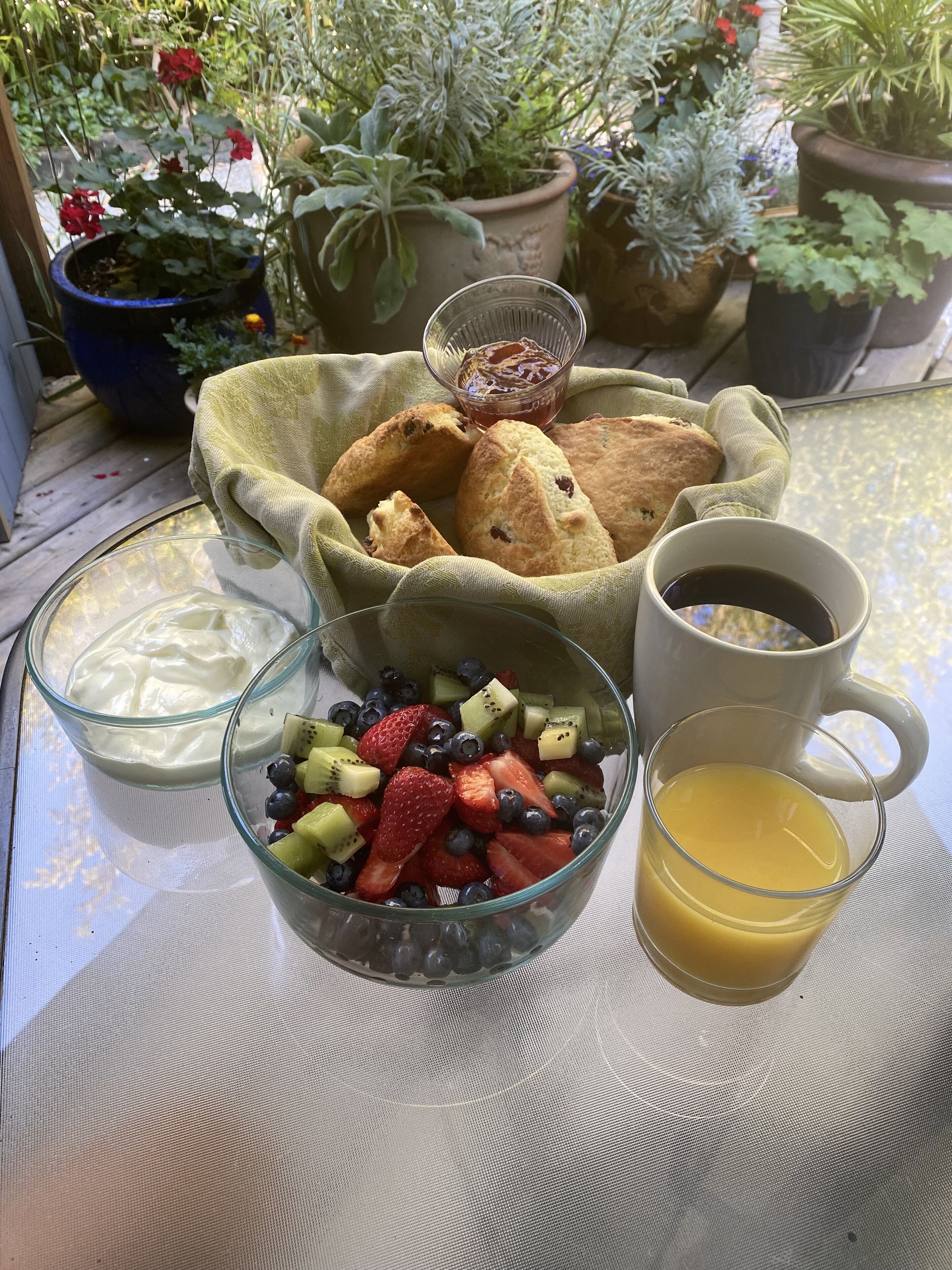 A breakfast table with a bowl of fruit, a glass of orange juice, a cup of coffee, and a basket of scones.