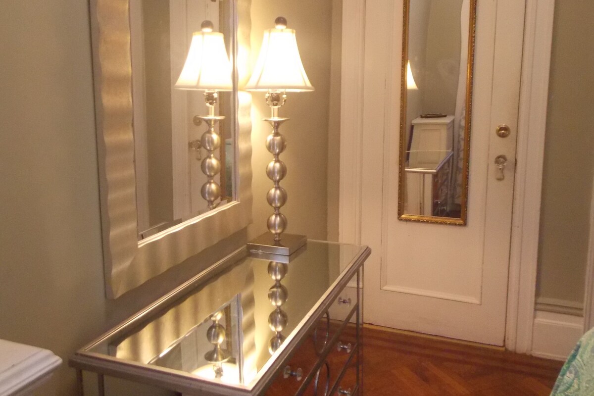 A bathroom with a large mirror, two wall-mounted lamps, a vanity with drawers, and a door with a mirror.