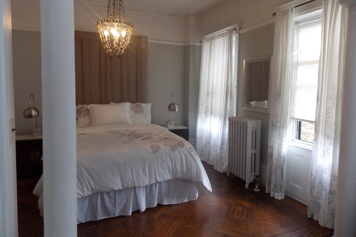 A bedroom with a large bed, a chandelier, a radiator, and a window with curtains.
