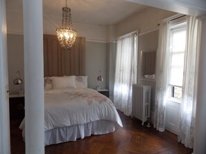 A bedroom with a large bed, a chandelier, a radiator, and a window with curtains. - Serenity at Home Guest House (Brooklyn)
