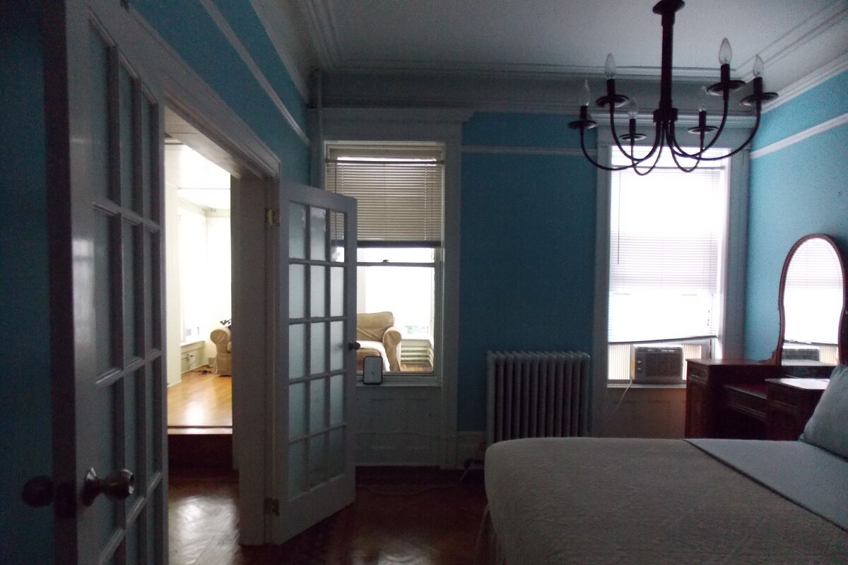 A bedroom with a bed, a dresser, a mirror, and a chandelier. There is a door leading to another room.