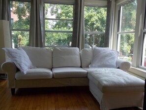 A white sectional sofa with a matching ottoman, positioned in front of a large window with multiple panes, offering a view of trees and a building outside. - Serenity at Home Guest House (Brooklyn)