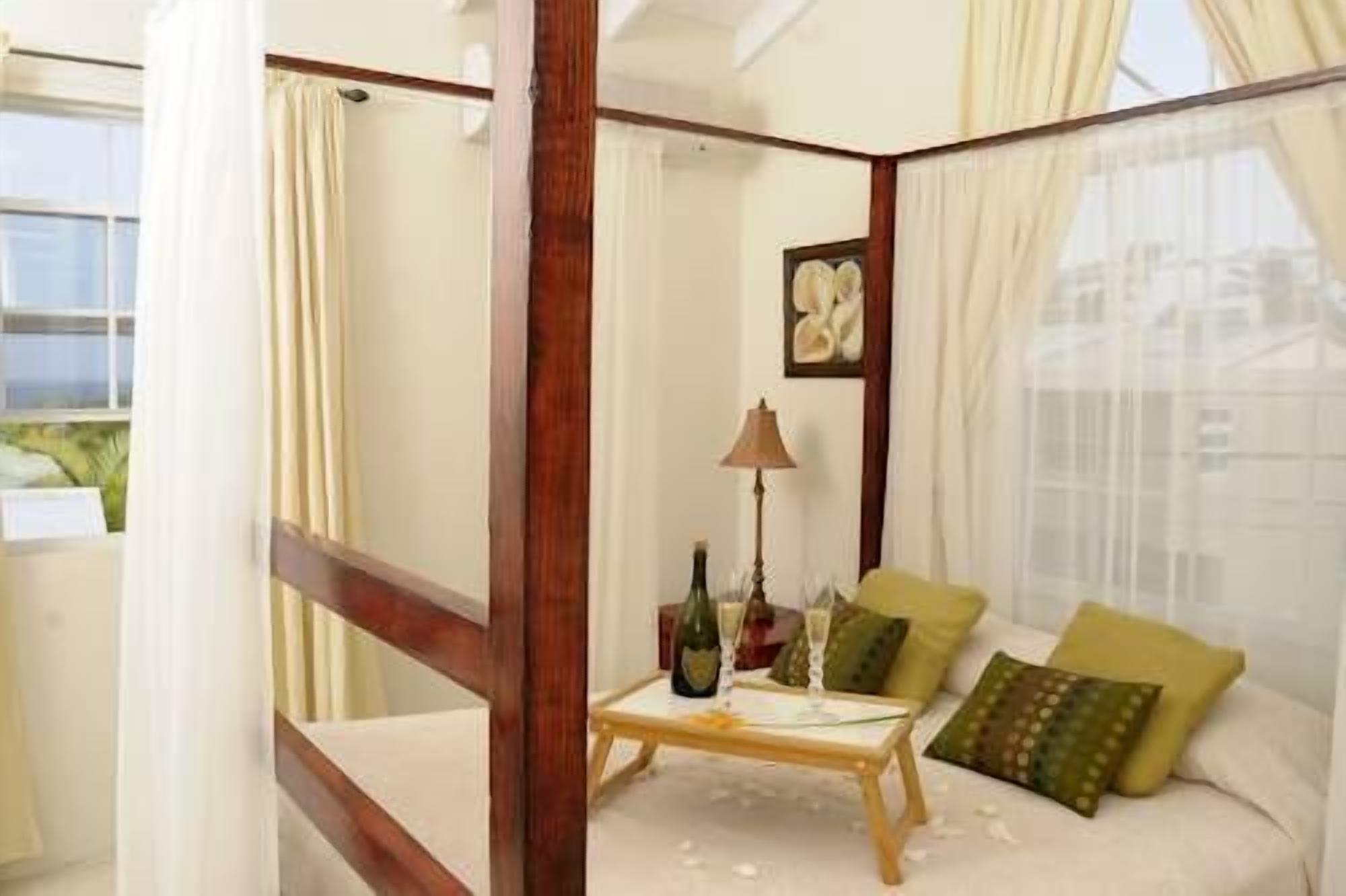 A bedroom with a four-poster bed, a small table with a bottle and glasses, and a cityscape view through sheer curtains.