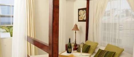 A bedroom with a four-poster bed, a small table with a bottle and glasses, and a cityscape view through sheer curtains.