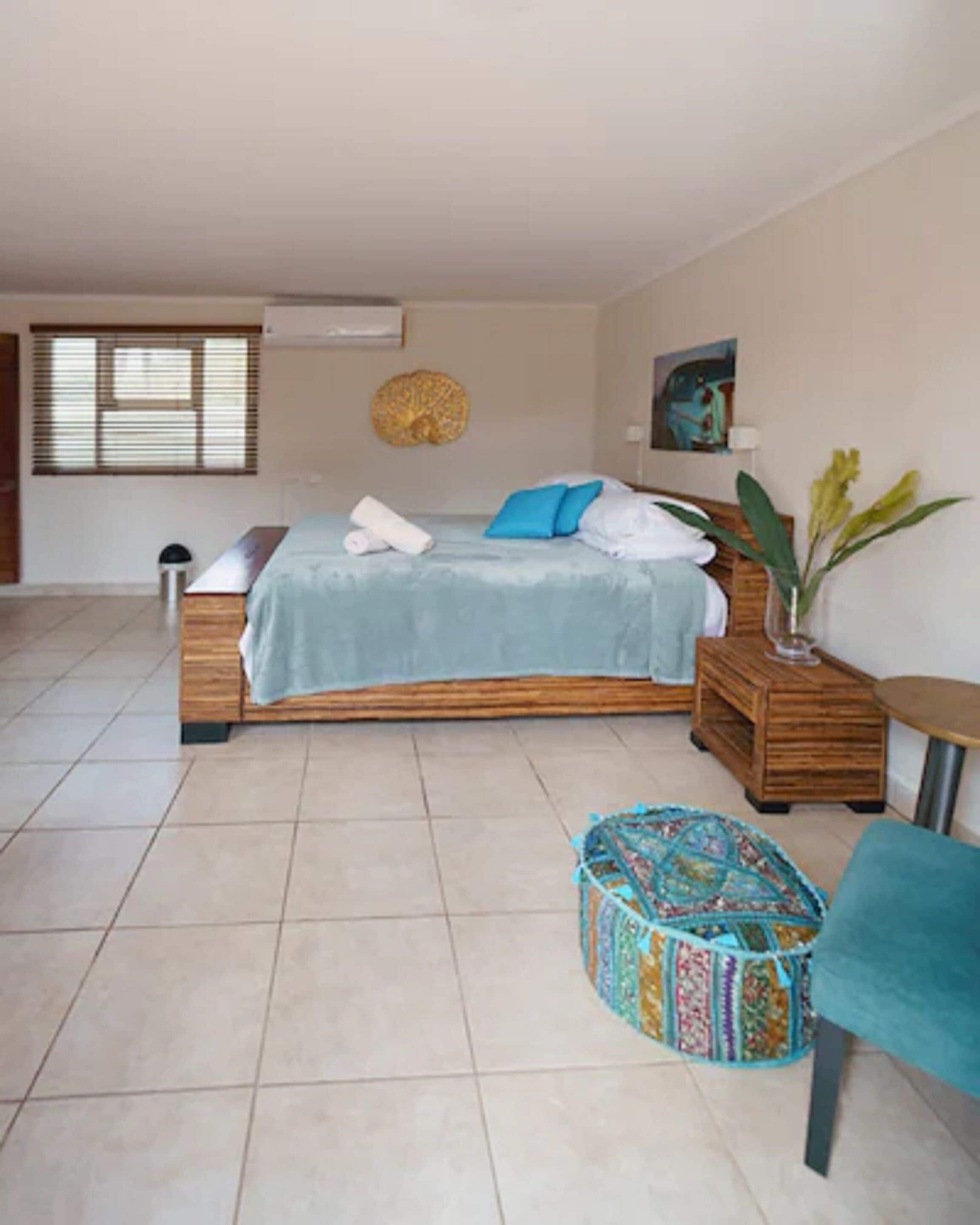 A bedroom with a bed, a bedside table, a small round table, a potted plant, and a colorful ottoman.
