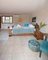 Un dormitorio con cama, mesita de noche, una pequeña mesa redonda, una planta en maceta y un otoman colorido.