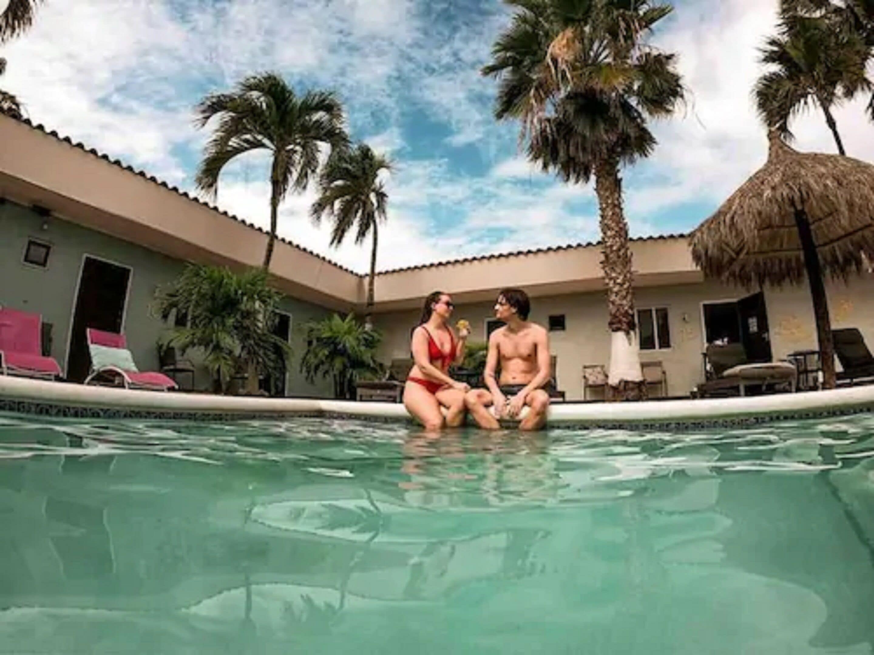 Un área de piscina con dos personas sentadas junto a ella, rodeada de palmeras y un edificio con techo de paja.