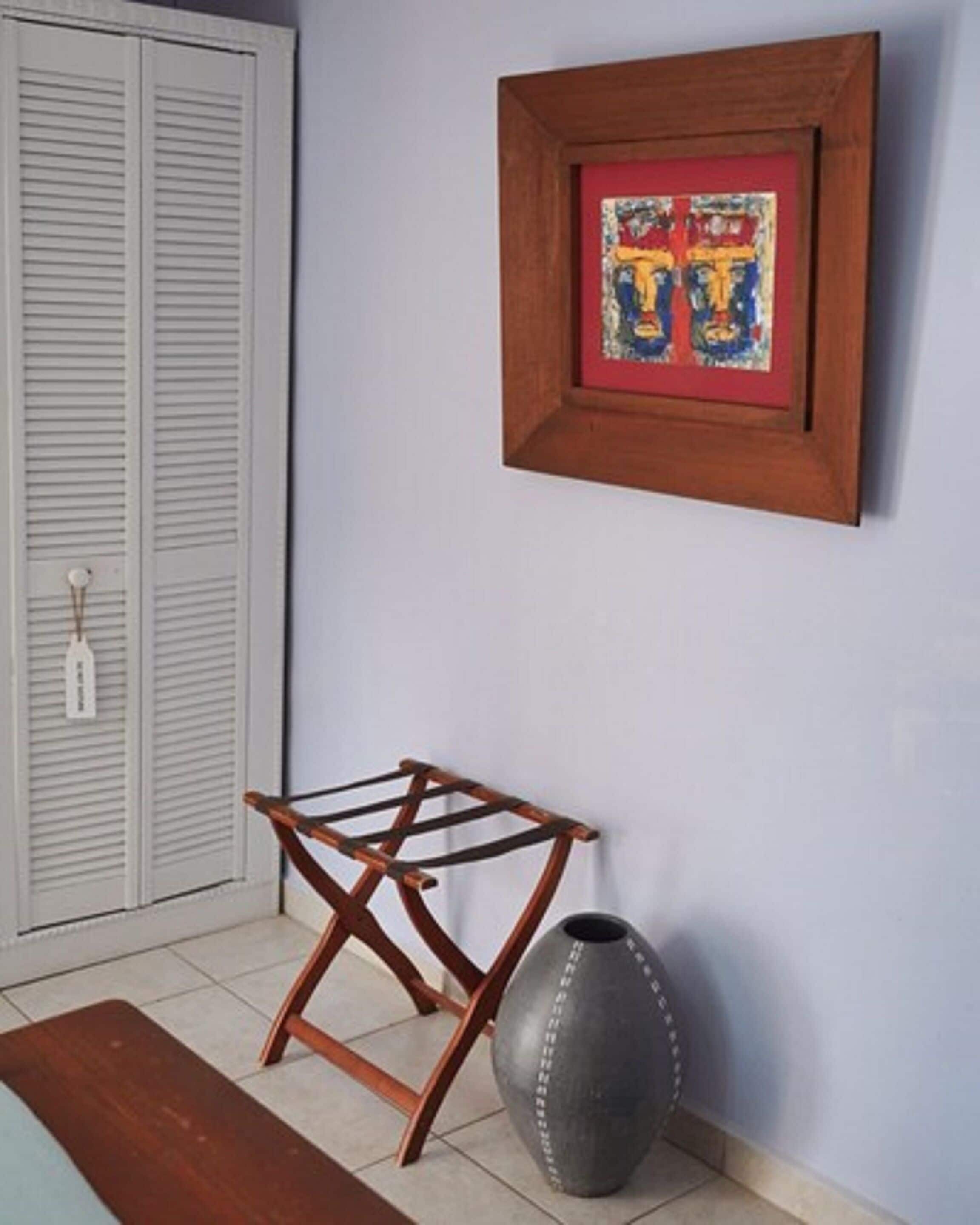 Una habitación con una silla plegable de madera, un jarrón gris y una obra de arte enmarcada en la pared.