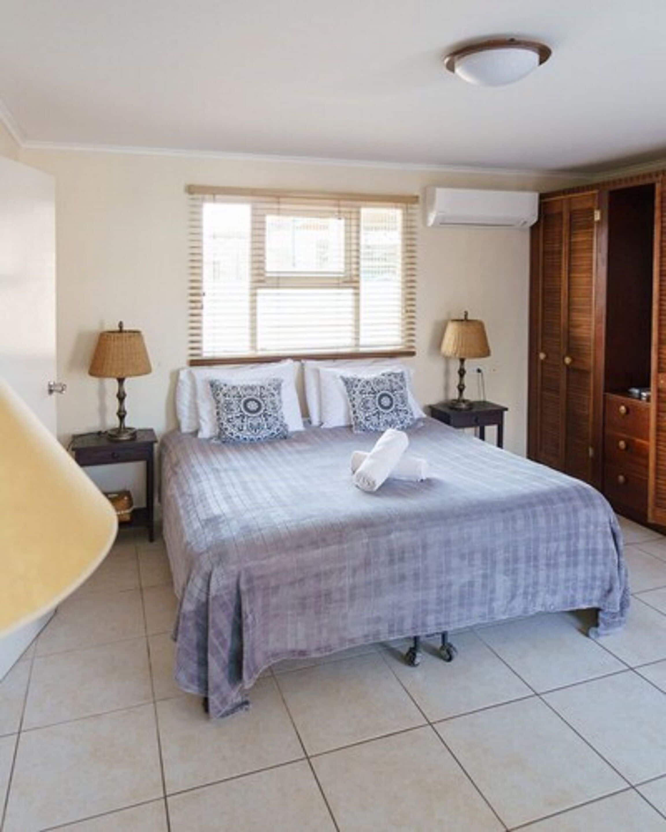 Een slaapkamer met een bed, twee nachtkastjes met lampen, een raam met jaloezieën, een kledingkast en een stoel.