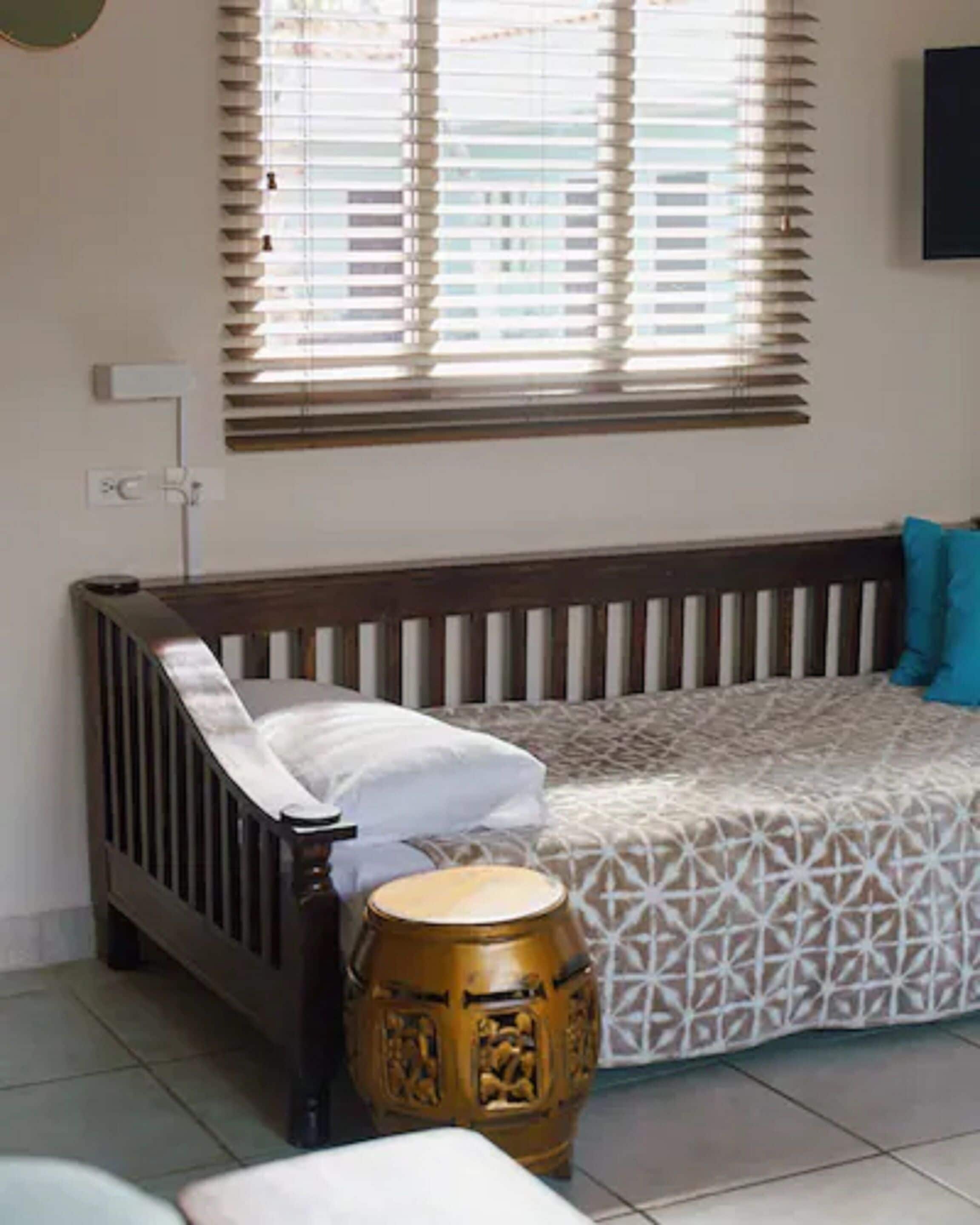 Quarto com berço de madeira, sofá estampado, um tambor e janela com persianas.