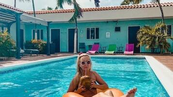 A swimming pool with a person on an orange float, surrounded by palm trees and a building with a tiled roof.