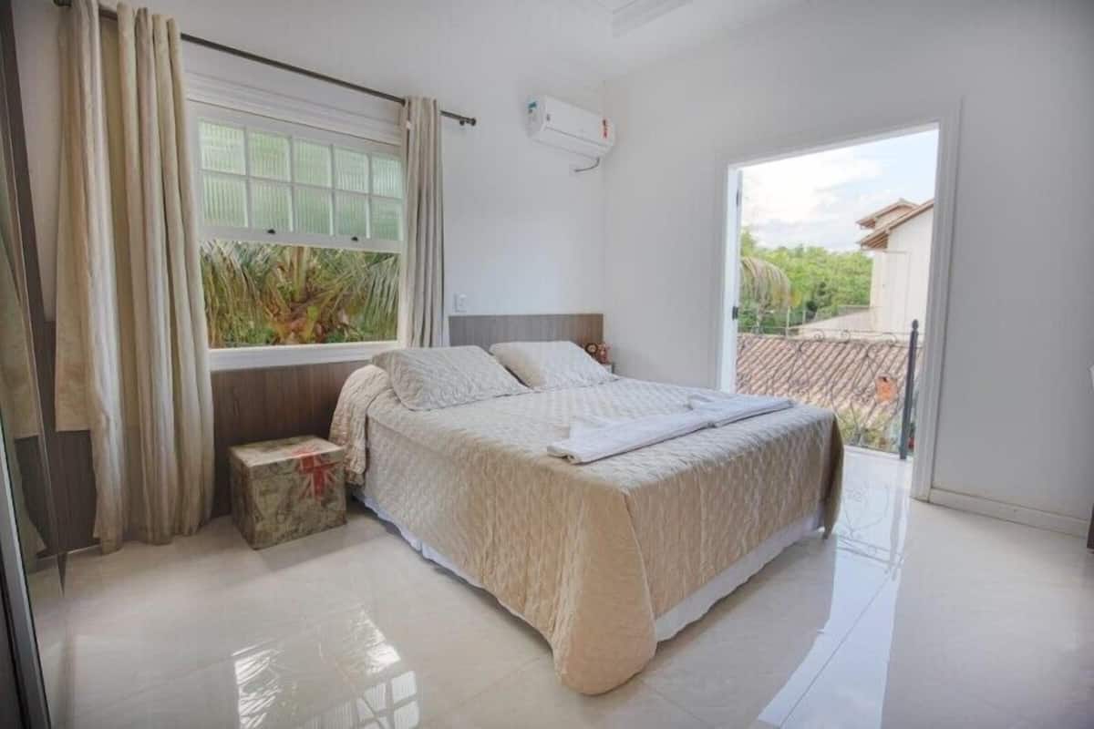 A bedroom with a bed, a window, an air conditioning unit, and a view of the outside.