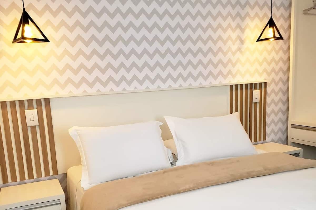 A hotel room with a bed, two bedside tables, a nightstand, and two pendant lights.