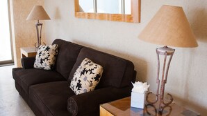 Una sala de estar con un sofá marrón oscuro, dos almohadones estampados, una mesa de centro de vidrio, una caja de pañuelos blancos y dos lámparas de pie.