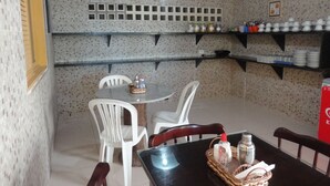 A dining area with a round table, white chairs, a clock, shelves with jars, and a red cooler. - Pousada Residence Oásis (Maragogi)