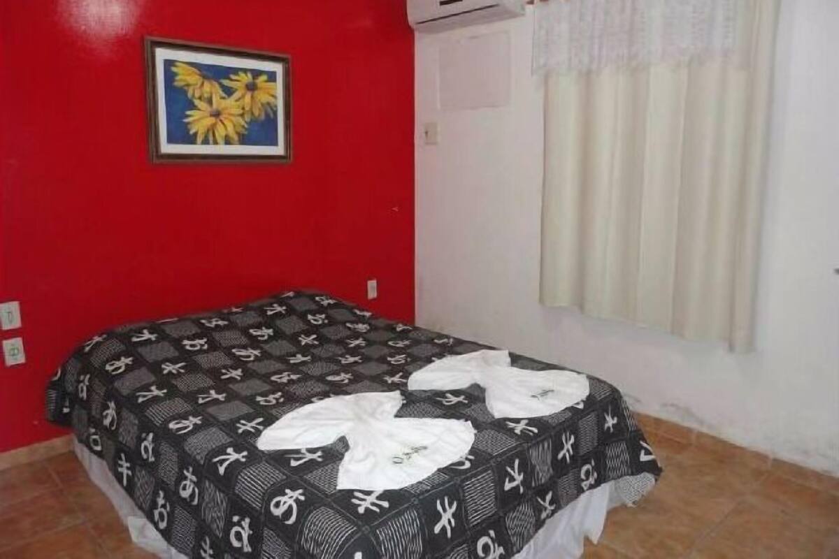 Ein Zimmer mit roter Wand, einem Bett mit gemustertem Überwurf, einem gerahmten Bild von Blumen und einem Fenster mit Vorhängen.
