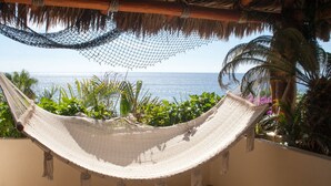Eine Hängematte auf einem Balkon mit Blick aufs Meer, umgeben von tropischen Pflanzen und einem Reetdach.