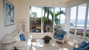 Ein Wohnzimmer mit einem weißen Sofa, einem blauen Sessel und einem Couchtisch. Ein großes Fenster bietet Blick auf Palmen und das Meer.