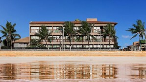 Un hôtel en bord de mer, avec plusieurs étages, des palmiers et une plage de sable au premier plan.