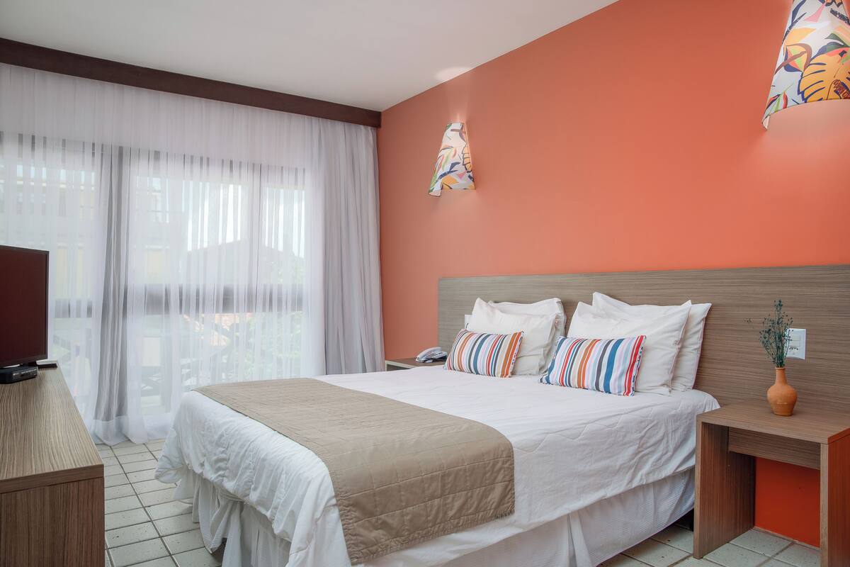 Habitación de hotel con una cama grande, ropa de cama blanca, cabecera de madera, mesita de noche con un jarrón y vistas a través de cortinas traslúcidas.