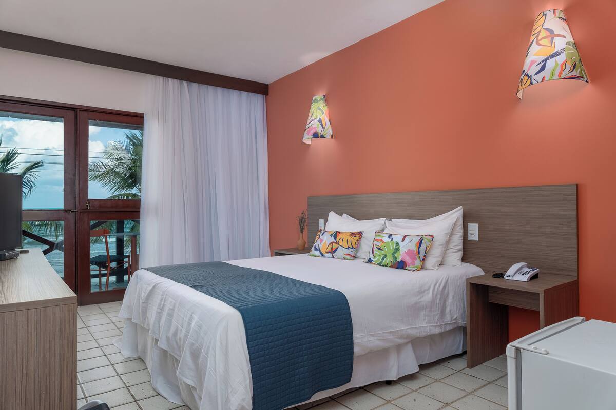 Habitación de hotel con cama, escritorio, silla, ventana con cortinas y vista a palmeras.