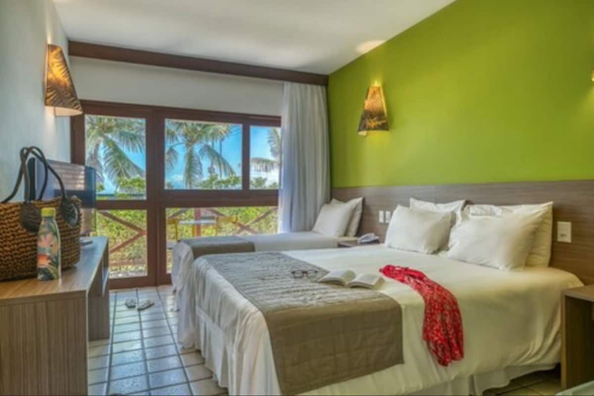 Habitación de hotel con una cama grande, un escritorio con lámpara y vistas a palmeras por la ventana.