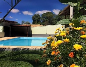 A swimming pool with a tiled deck, surrounded by a grassy area and yellow flowers. - Pousada da Lapa (Vespasiano)