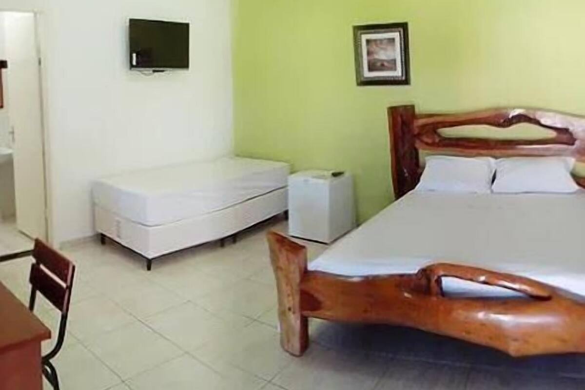 Quarto com cama de madeira, colchão branco, pequena mesa branca, uma cadeira, televisão na parede e um porta-retrato.