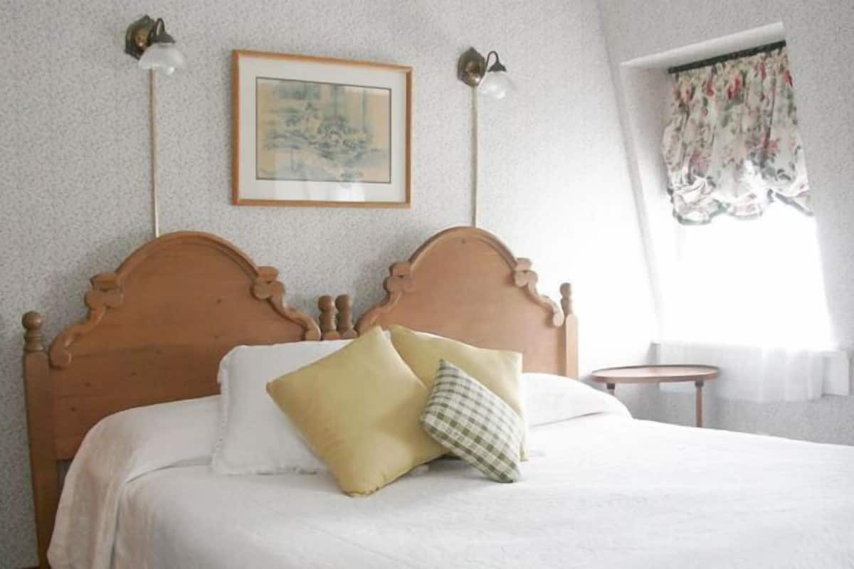 A bedroom with a wooden bed, two wall-mounted lamps, a framed picture, and a window with floral curtains.