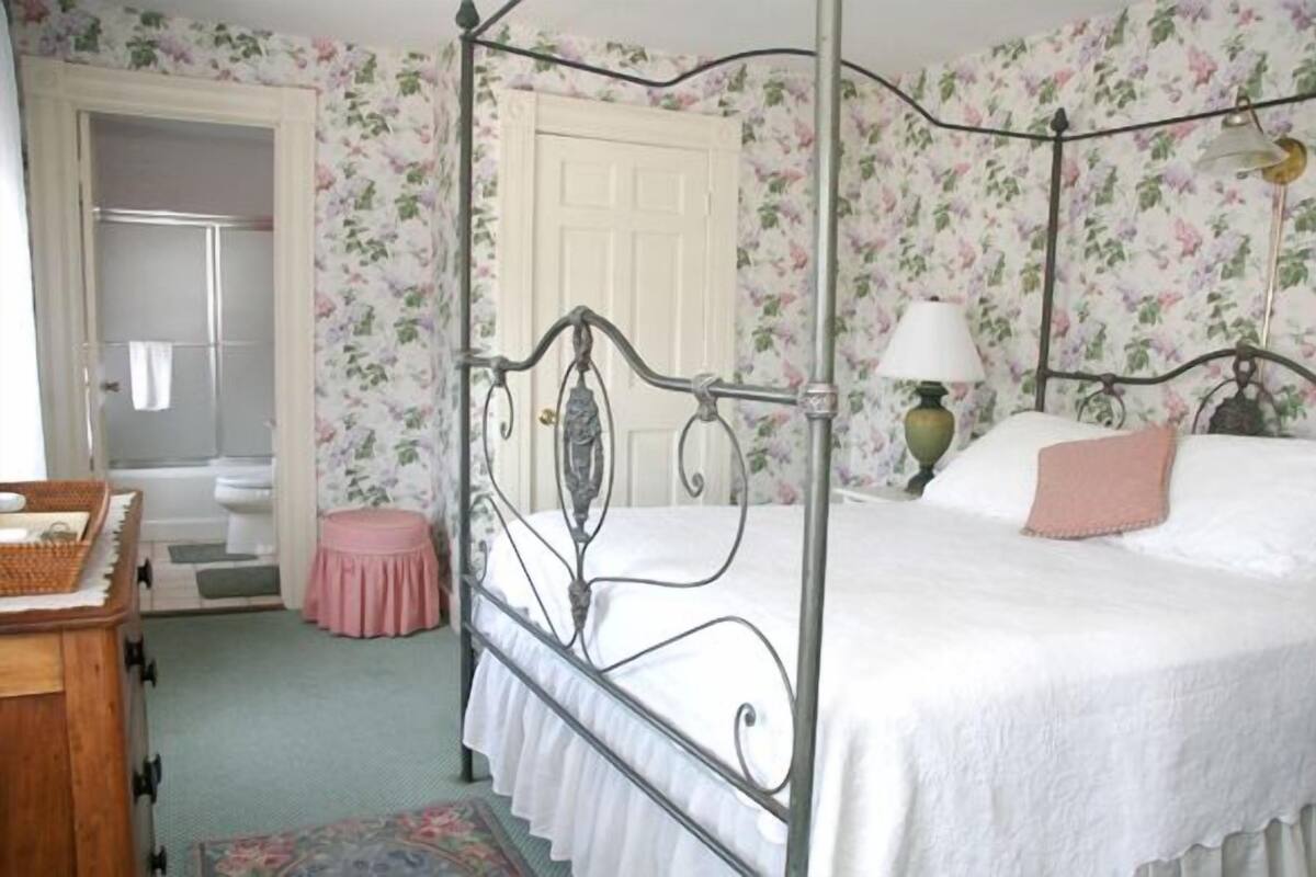 A bedroom with a four-poster bed, a small stool, a wooden dresser, and a door leading to a bathroom.