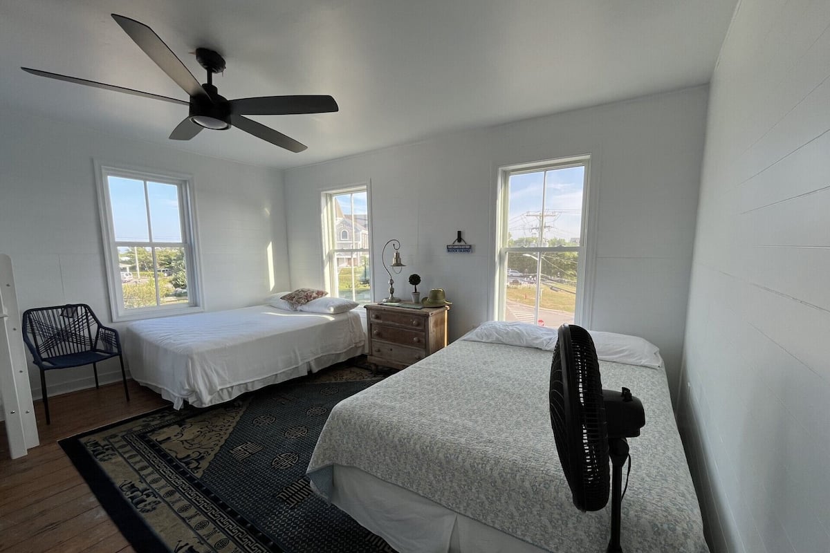 A bedroom with two beds, a ceiling fan, a standing fan, a chair, and a window with a view of a building.