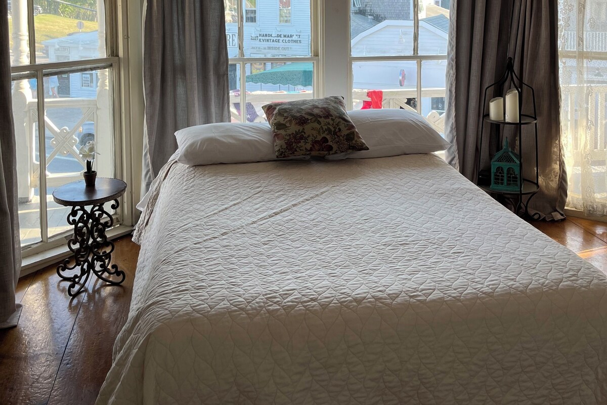 Un dormitorio con una cama grande, una ventana con cortinas, una mesita y una mesita de noche con una lámpara.
