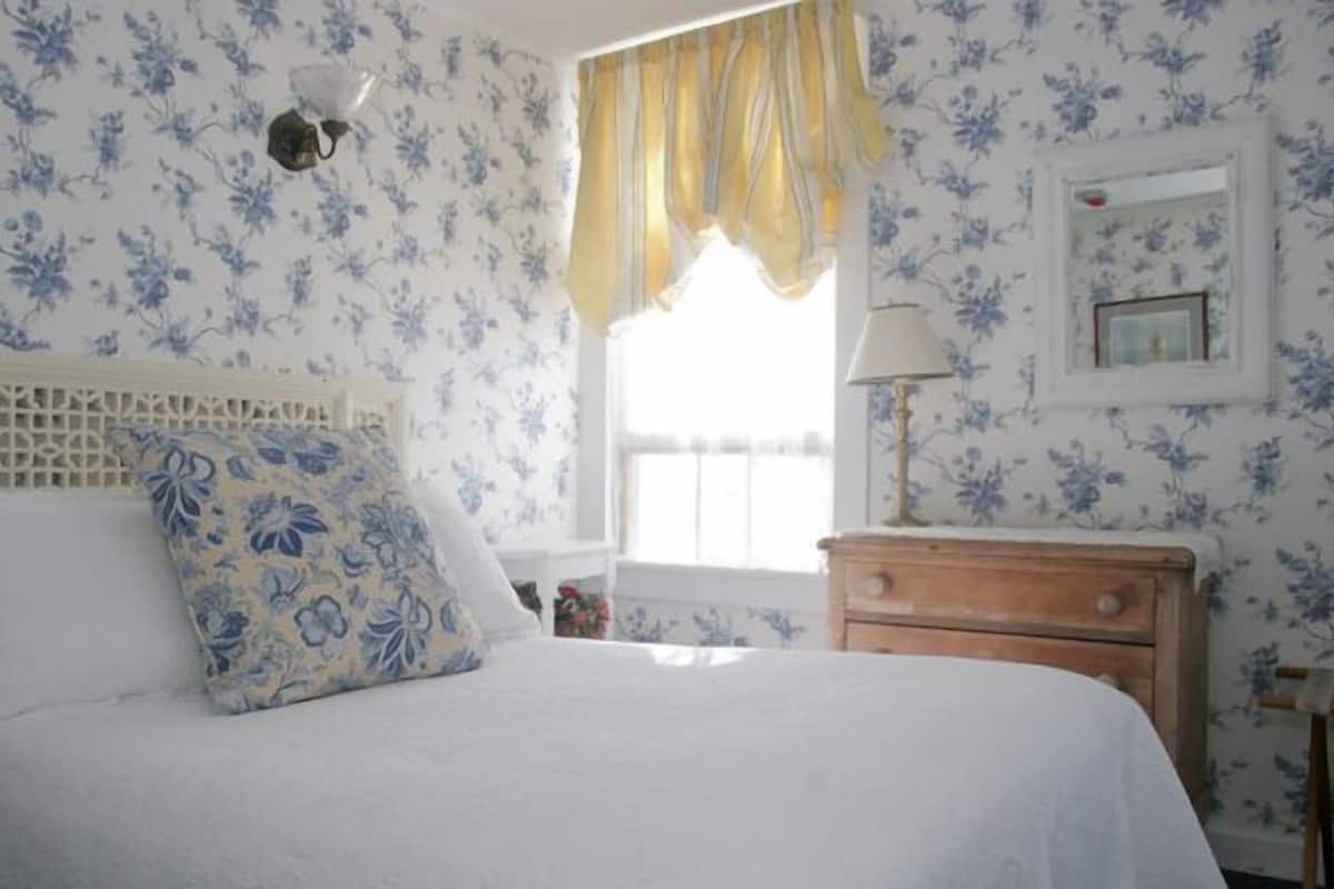 A bedroom with a bed, a nightstand, a lamp, a window with curtains, and floral wallpaper.