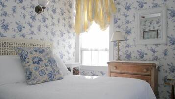 A bedroom with a bed, a nightstand, a lamp, a window with curtains, and floral wallpaper.