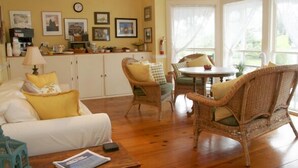 A cozy living room with a sofa, wicker chairs, a dining table, and a kitchen area with cabinets and a clock. - Inn at Old Harbor (Block Island)
