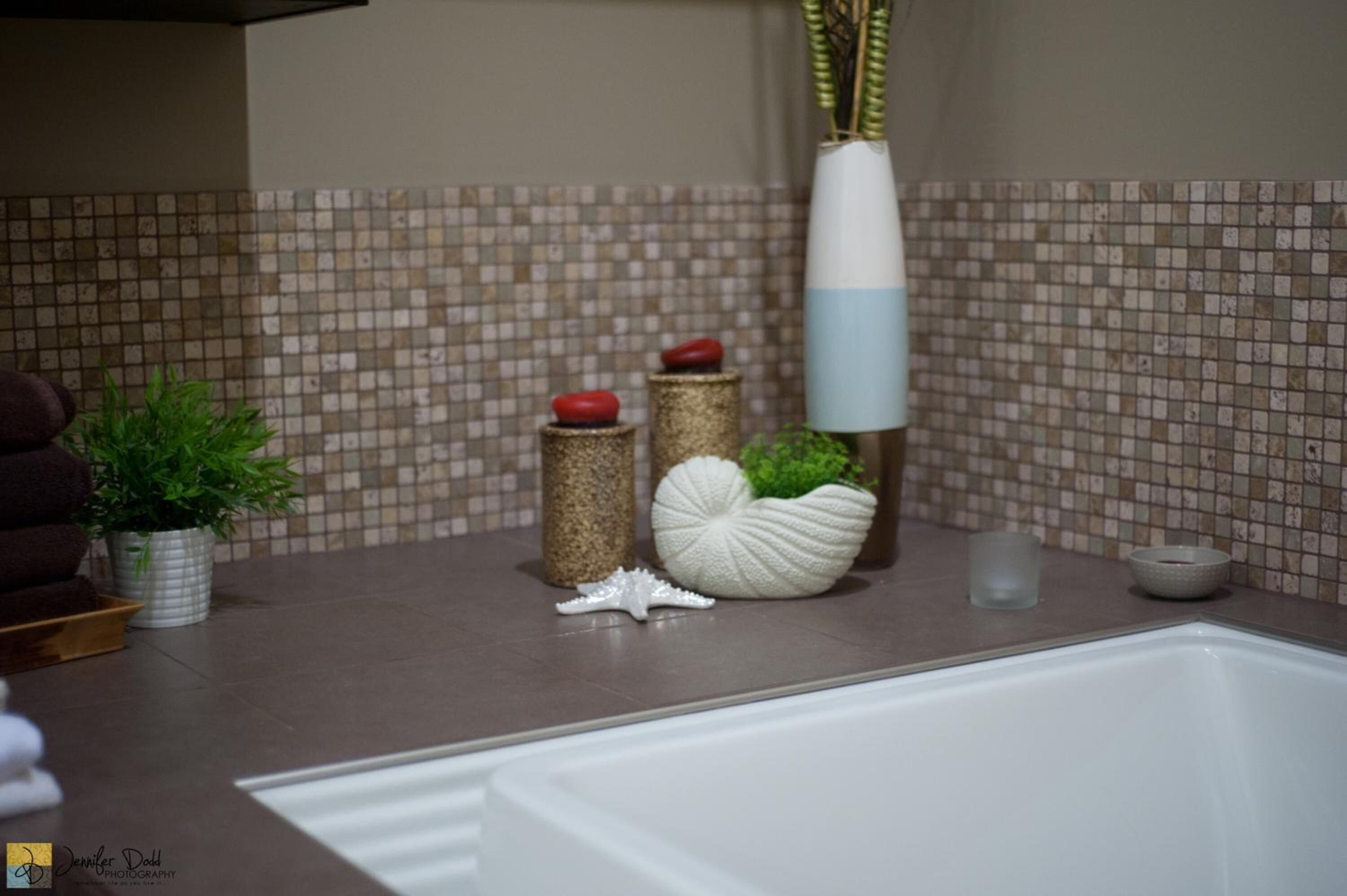 Um banheiro com painel de azulejos, uma banheira e vários objetos decorativos, incluindo uma planta em vaso, um recipiente branco em formato de concha e um vaso alto com folhagem verde.
