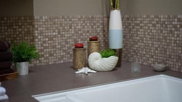 Um banheiro com painel de azulejos, uma banheira e vários objetos decorativos, incluindo uma planta em vaso, um recipiente branco em formato de concha e um vaso alto com folhagem verde.