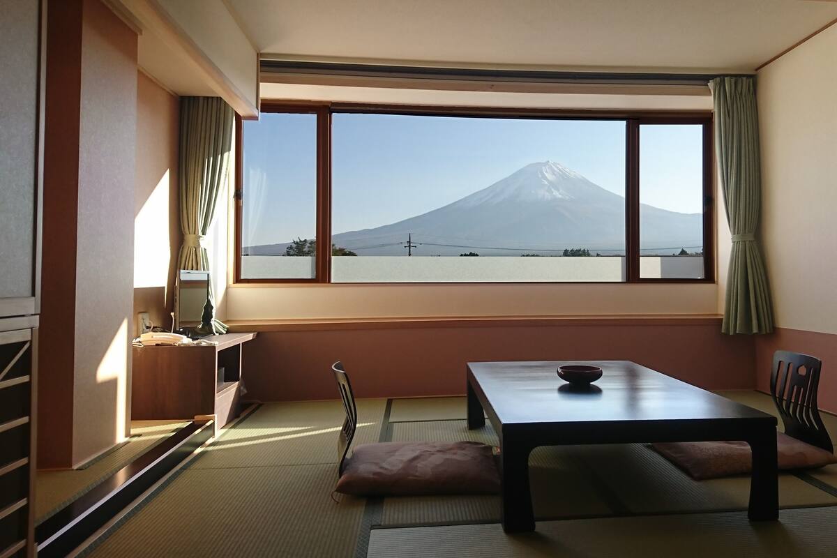 Habitació amb terra de tatami, una taula baixa amb un bol, una cadira i vista a una muntanya.
