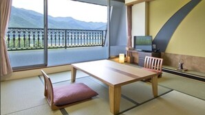 A traditional Japanese room with tatami flooring, a low table, a cushion, a television, and a view of mountains through large windows. - Lakeland Hotel Mizunosato (Fujikawaguchiko)