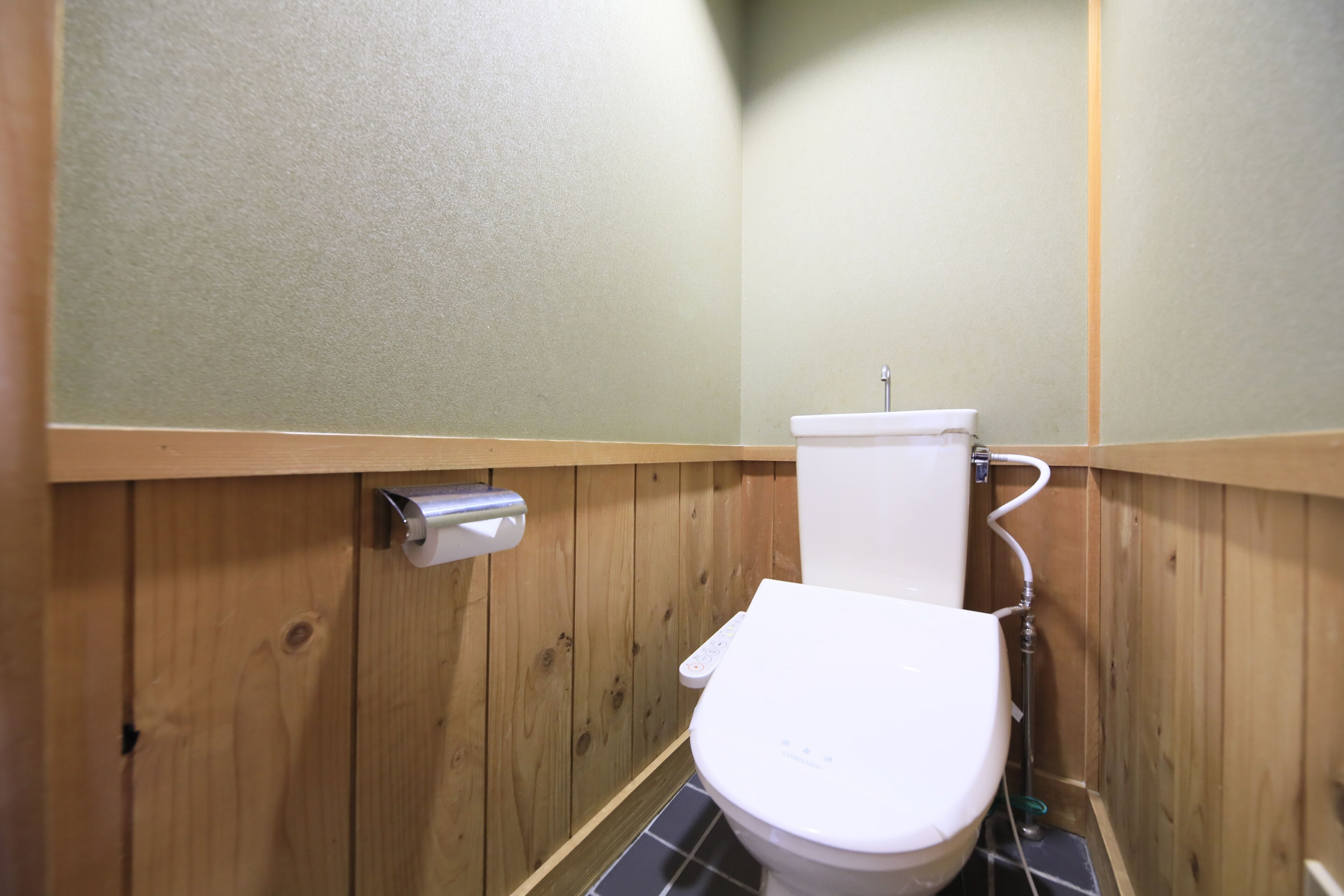 A small, wooden-paneled bathroom with a white toilet, a hand dryer, and a visible pipe.