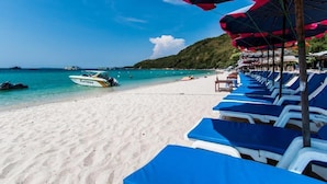 A beach with rows of blue lounge chairs and umbrellas, a boat named "KATNISA" on the sand, and a clear blue sea with boats in the distance. - Suntosa Resort (Koh Lan)