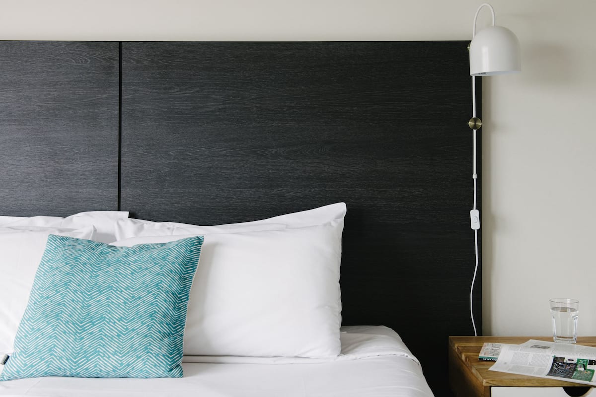 A neatly made bed with white and teal pillows, a dark headboard, a bedside table with a glass of water, and a lamp.