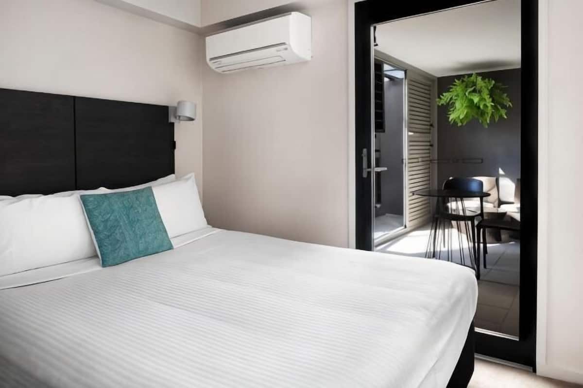 A modern hotel room with a large bed, a dark headboard, and a view of the outdoor seating area through an open door.