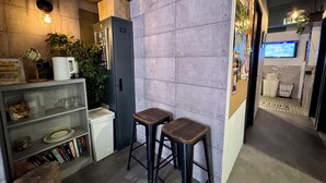 A compact kitchen area with a coffee machine, a small refrigerator, and a shelf with books and a kettle. There are two stools and a potted plant. - Urban Pack - Hostel (Kowloon)