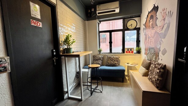A small, cozy room with a black door, a small table with a plant, a blue sofa, and a wall clock. - Urban Pack - Hostel (Kowloon)